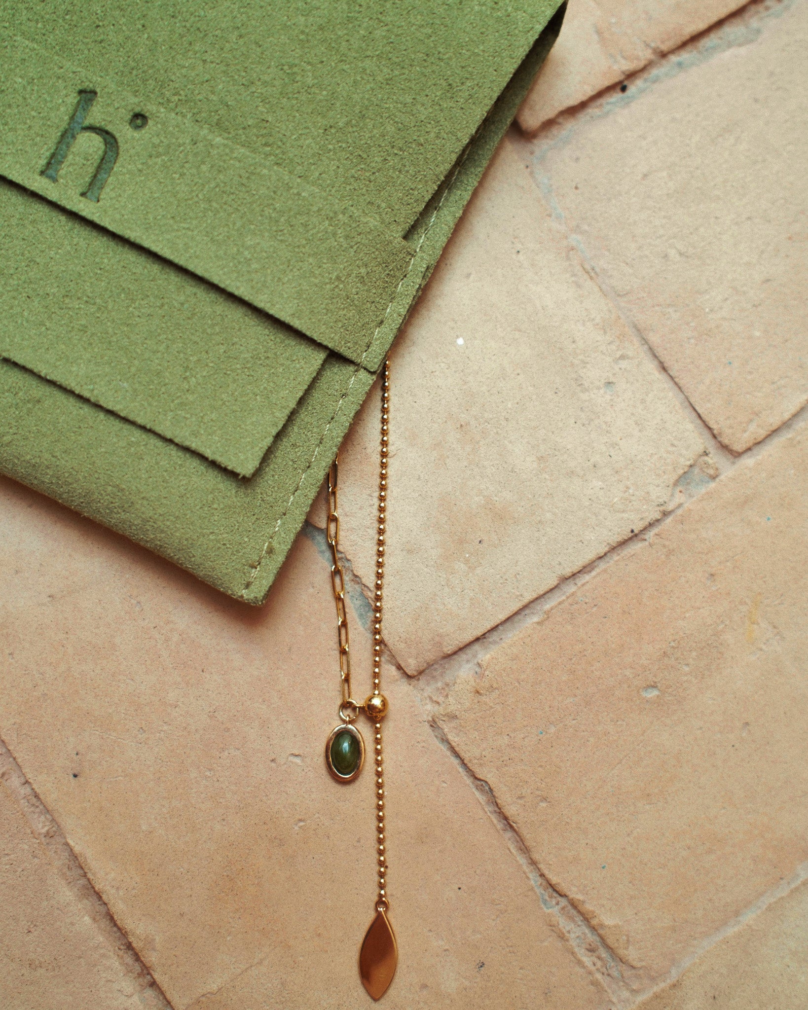Gold necklace with a pendant on a textured stone surface next to green packaging with 'h' heiam branding.
